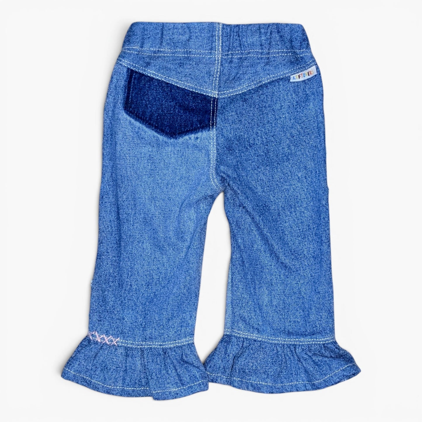 Blue denim pants for toddlers, featuring a back pocket with a darker blue patch, white contrast stitching, and ruffled hems.