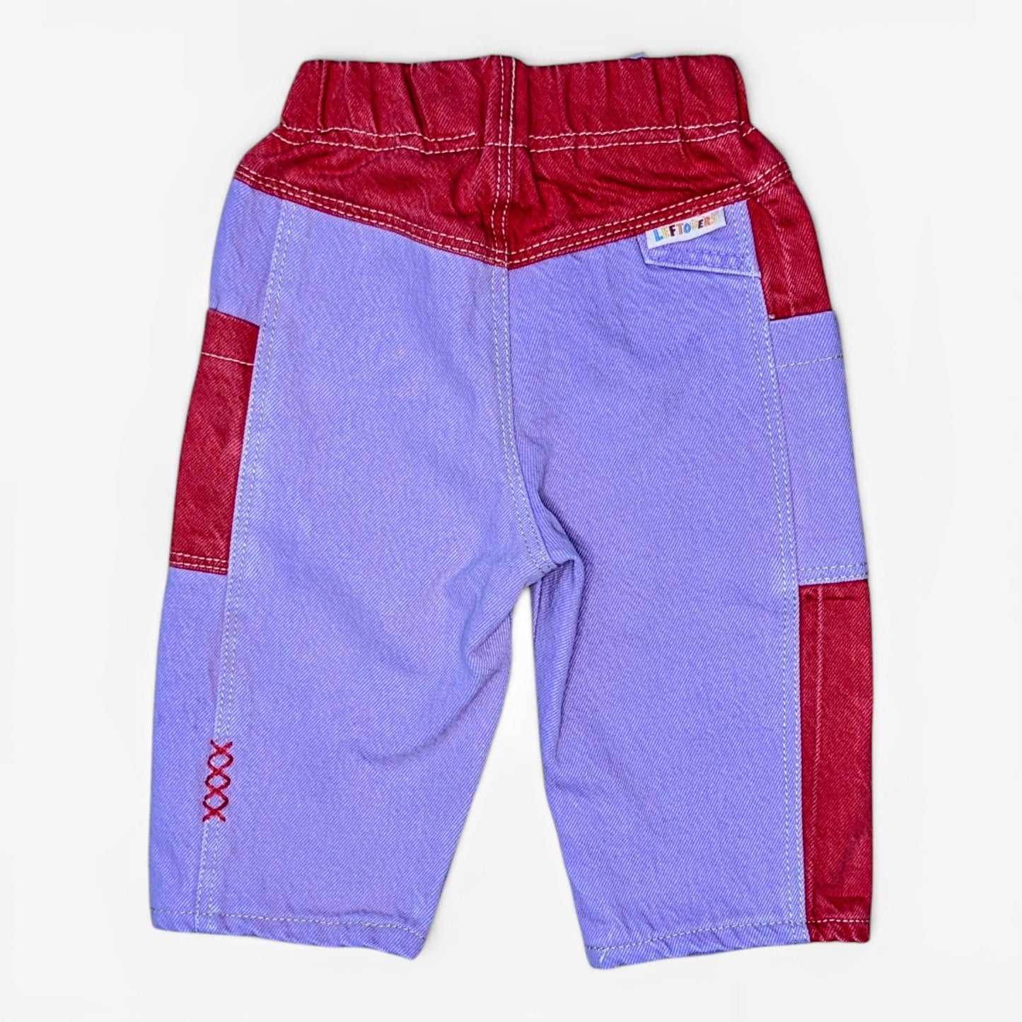 A pair of children's patchwork pants shown from the back, featuring a lavender denim-like body with red contrast panels at the waistband, side pockets, and lower leg seams.
