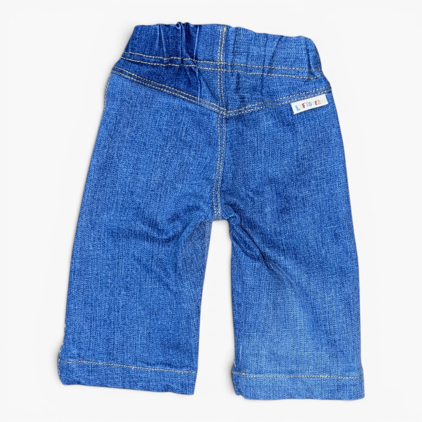 A pair of blue denim jeans for toddlers, shown from the back on a light off-white background. The jeans feature an elastic waistband, classic denim stitching, and a small white brand label on the upper right side.