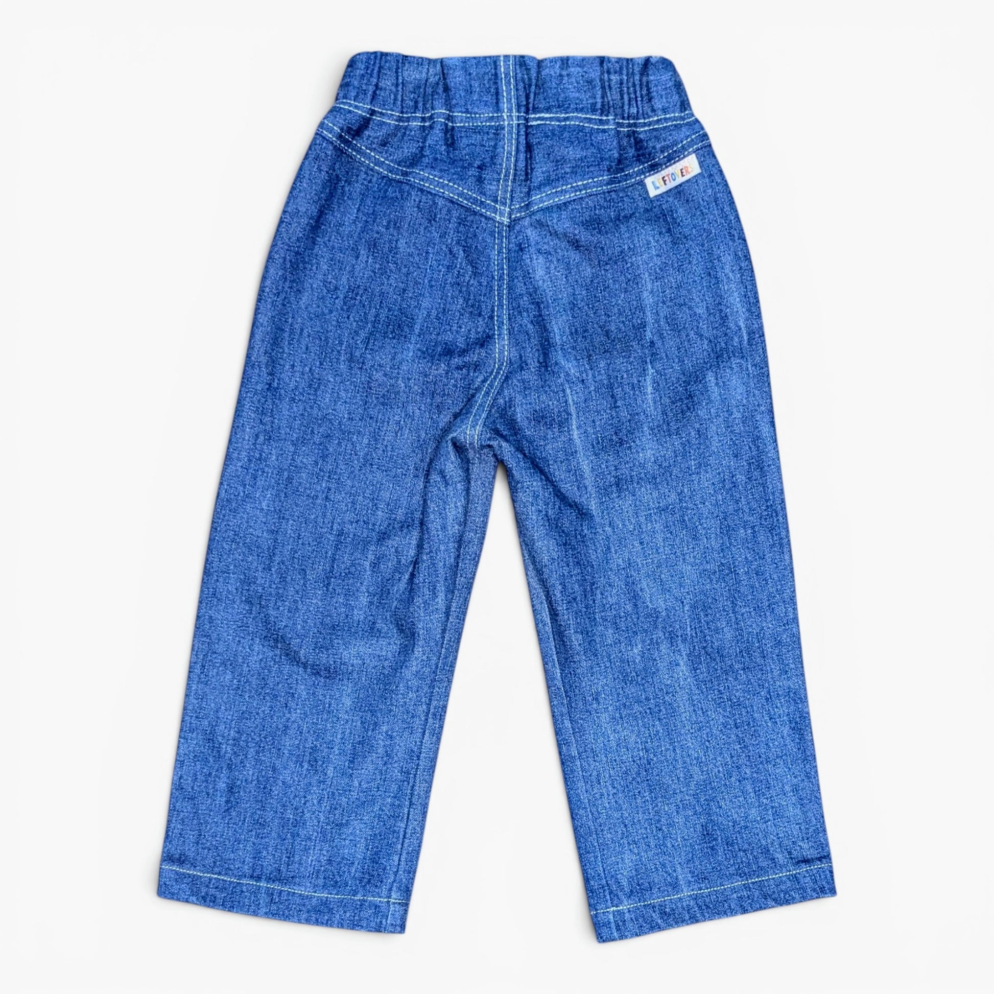 A pair of blue denim pants for toddlers, shown from the back on a light off-white background. The back features a wide elastic waistband, double-stitched seams, and a small white brand label on the upper right side.