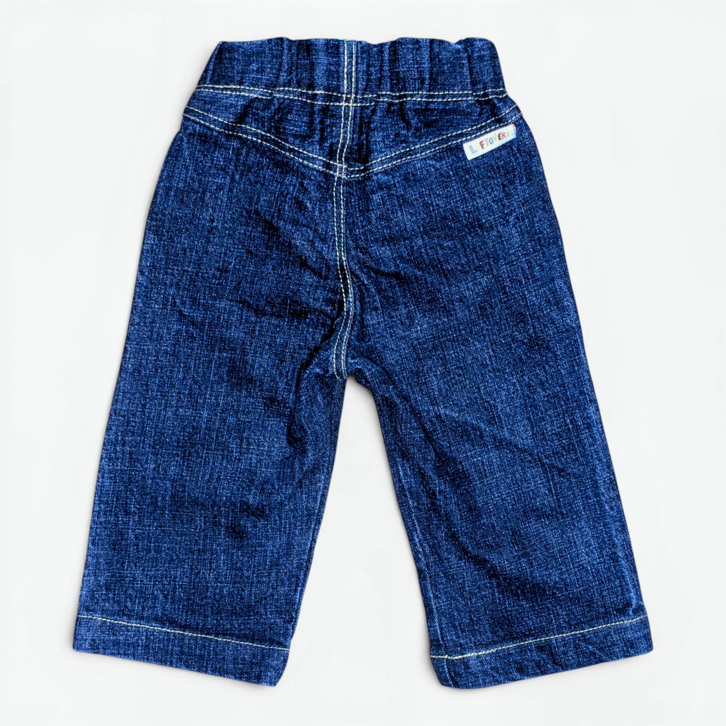 A pair of toddler denim pants in a medium blue wash, laid flat against a light off-white background. The pants feature a wide elastic waistband, contrast white stitching, and a small white brand label on the upper right hip.