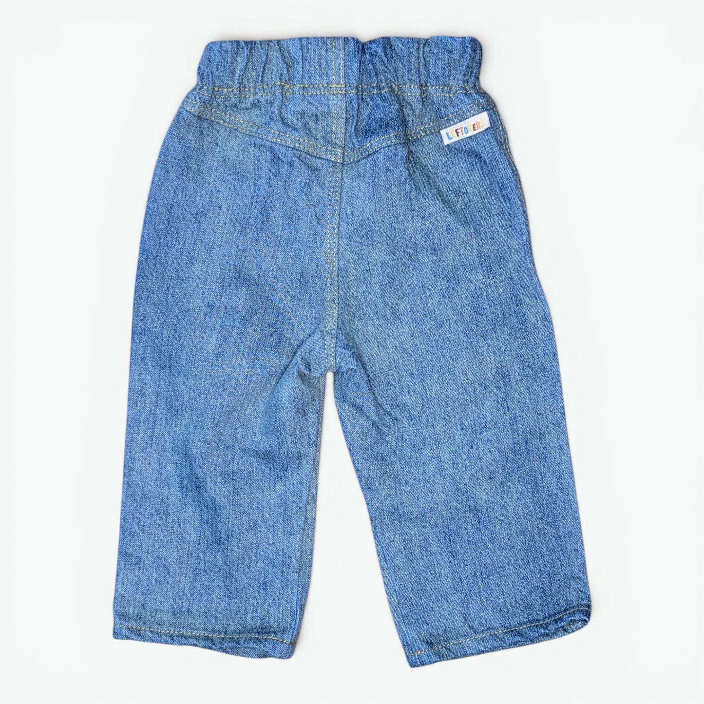 A pair of blue denim pants for toddlers, shown from the back on a light off-white background. The back features an elastic waistband, a central seam, and a small white brand tag on the upper right side.