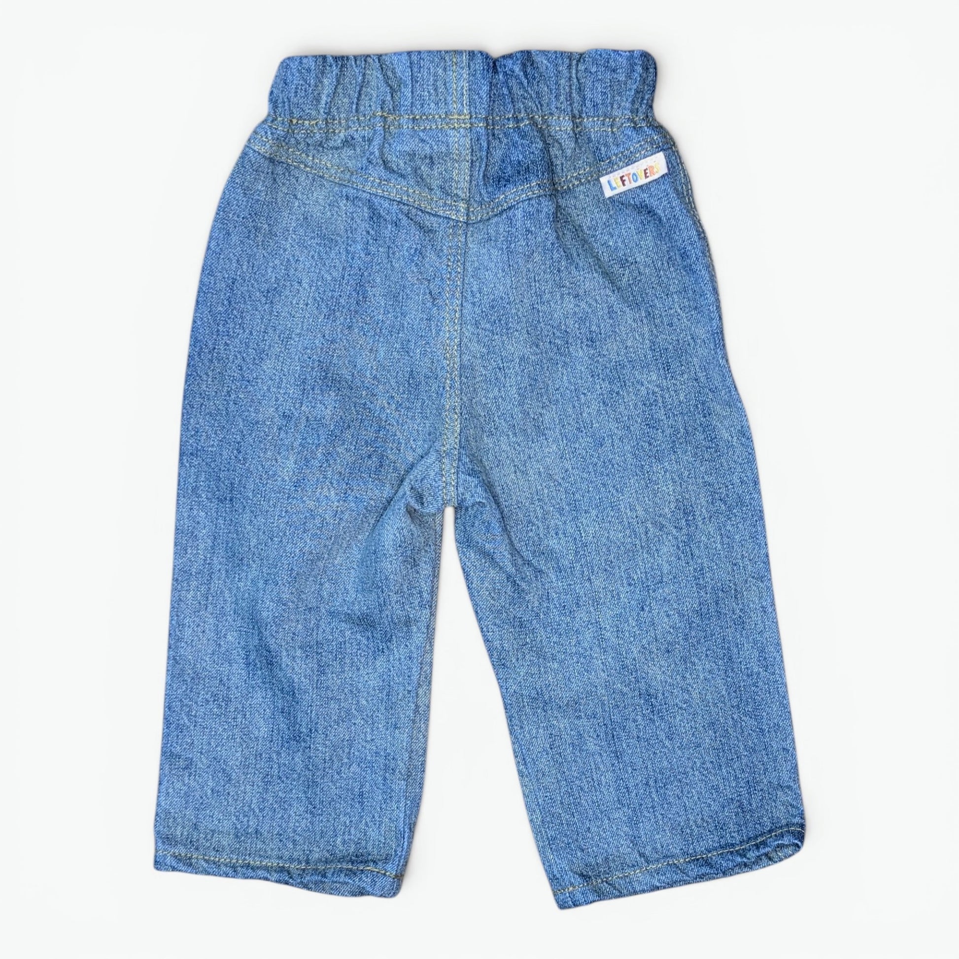 A pair of blue denim pants for toddlers, shown from the back on a light off-white background. The back features an elastic waistband, a central seam, and a small white brand tag on the upper right side.
