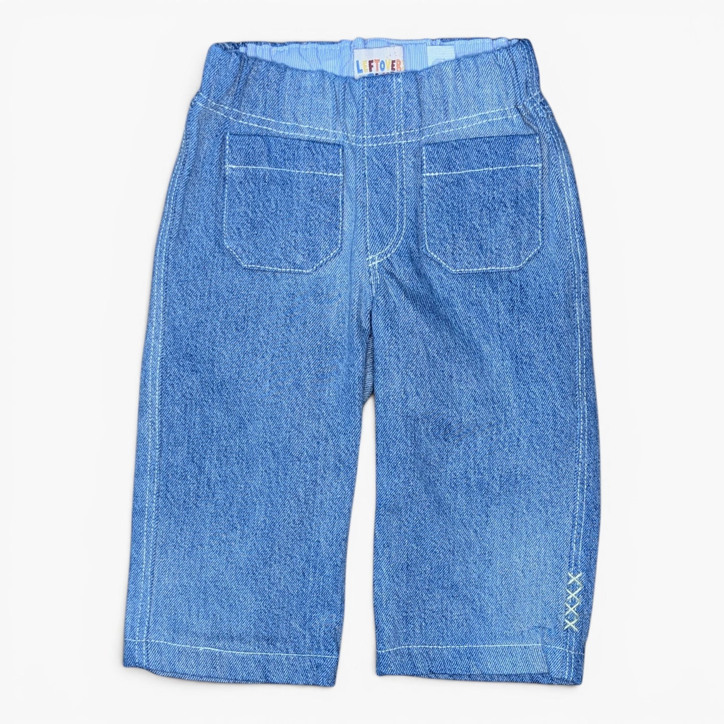 A pair of blue denim cropped pants shown from the back, featuring two large patch pockets, a wide elastic waistband, and decorative white stitching on the lower leg.