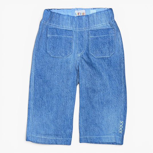 A pair of blue denim cropped pants shown from the back, featuring two large patch pockets, a wide elastic waistband, and decorative white stitching on the lower leg.