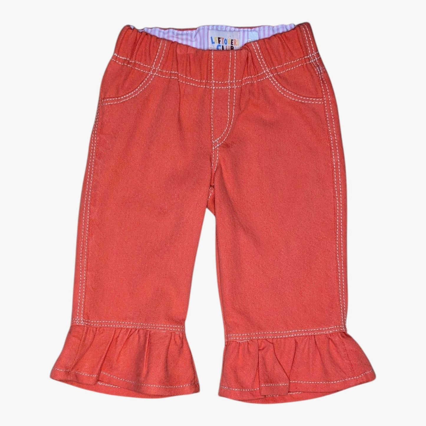 A pair of toddler's denim pants in a muted red-orange color, featuring white contrast stitching, and ruffled hems, shown against a plain white background.