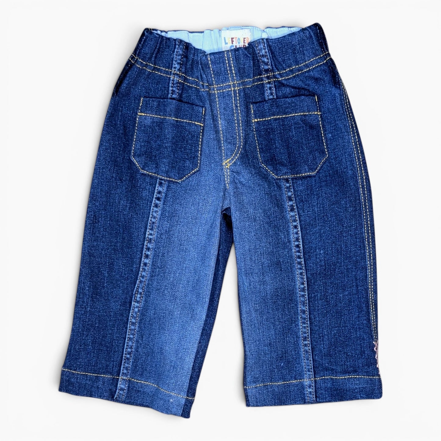 A pair of blue denim pants shown from the back, featuring two large patch pockets, contrast yellow stitching, and a light blue inner waistband, laid flat on a white background.