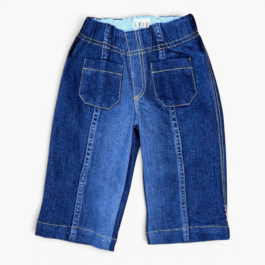 A pair of blue denim pants shown from the back, featuring two large patch pockets, contrast yellow stitching, and a light blue inner waistband, laid flat on a white background.