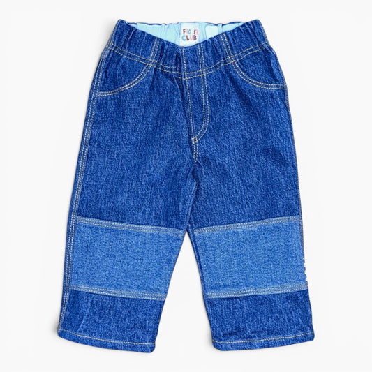 A pair of toddler denim pants in a blue wash, featuring horizontal patchwork panels on the lower legs and contrast light blue lining at the elastic waistband, shown flat on a white background.