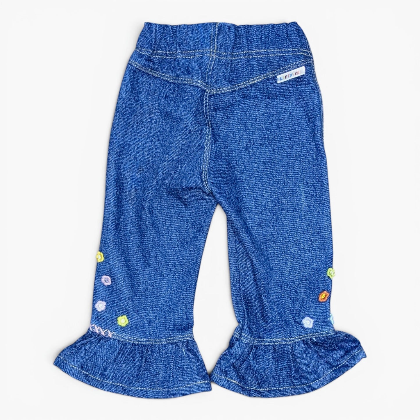 Blue denim pants for toddlers, featuring a ruffled hem and colorful embroidered accents on the legs, shown against a plain white background.