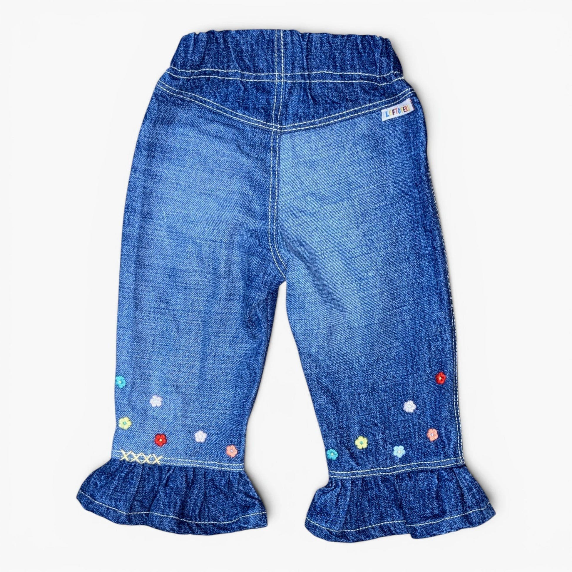 Blue denim toddler pants with a ruffled hem and colorful embroidered flowers, shown from the back on a light gray background.