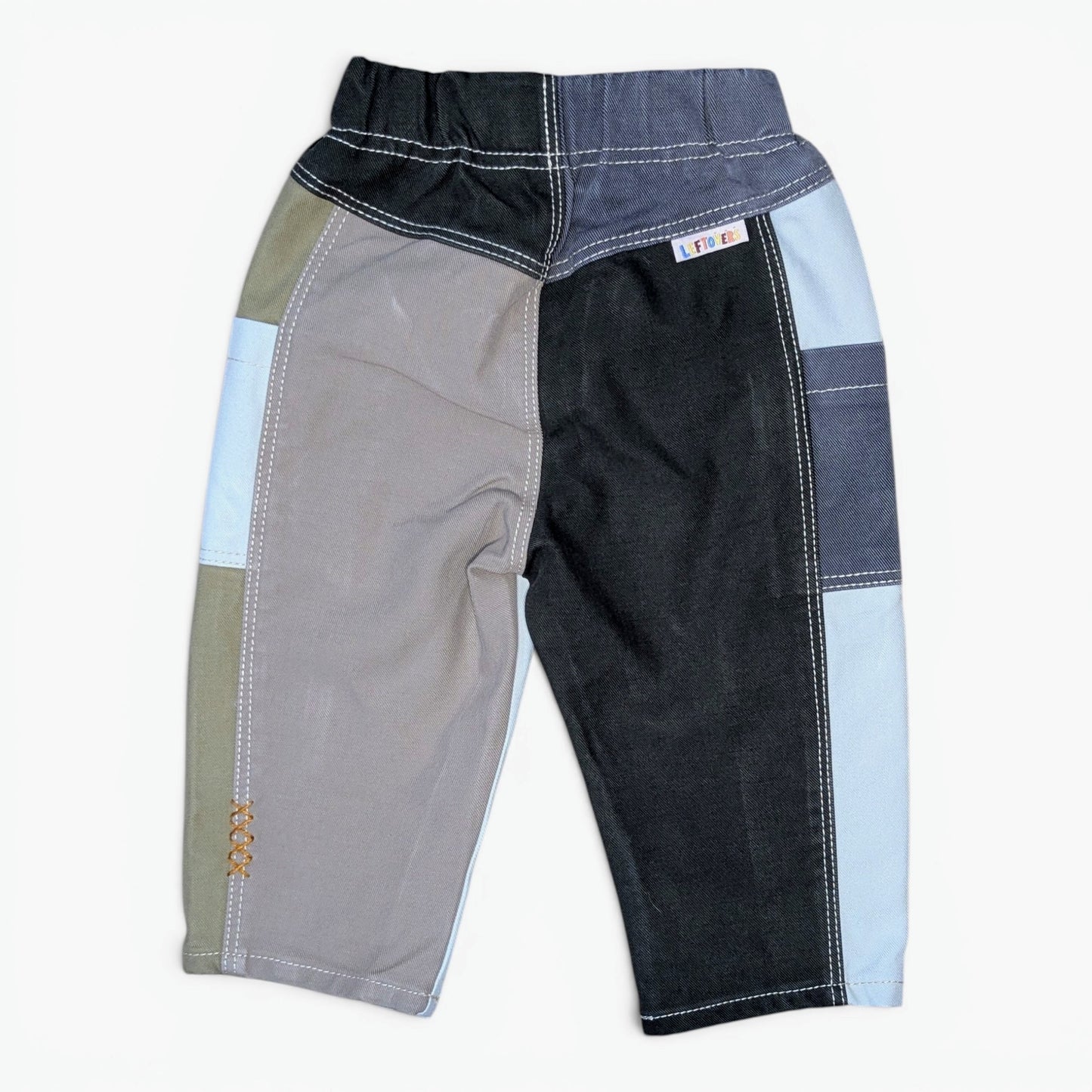 A pair of patchwork denim pants for kids, featuring color-blocked panels of black, gray, light blue, and olive green with contrast white stitching, shown flat on a white background.