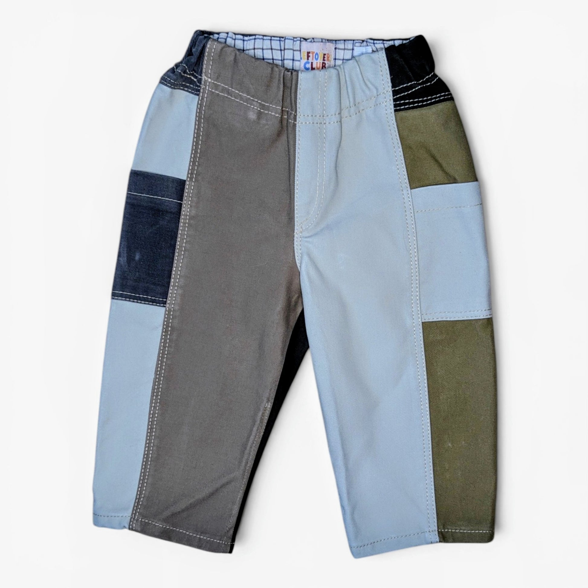 A pair of patchwork pants for kids, featuring color-blocked panels of gray, light blue, dark blue denim, olive green, and black, laid flat against a light off-white background.