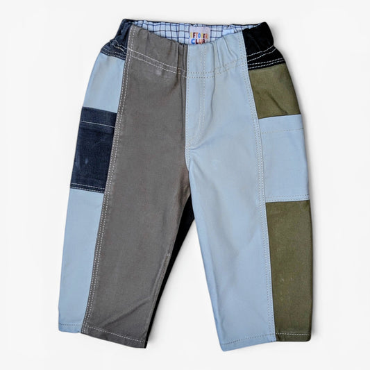 A pair of patchwork pants for kids, featuring color-blocked panels of gray, light blue, dark blue denim, olive green, and black, laid flat against a light off-white background.