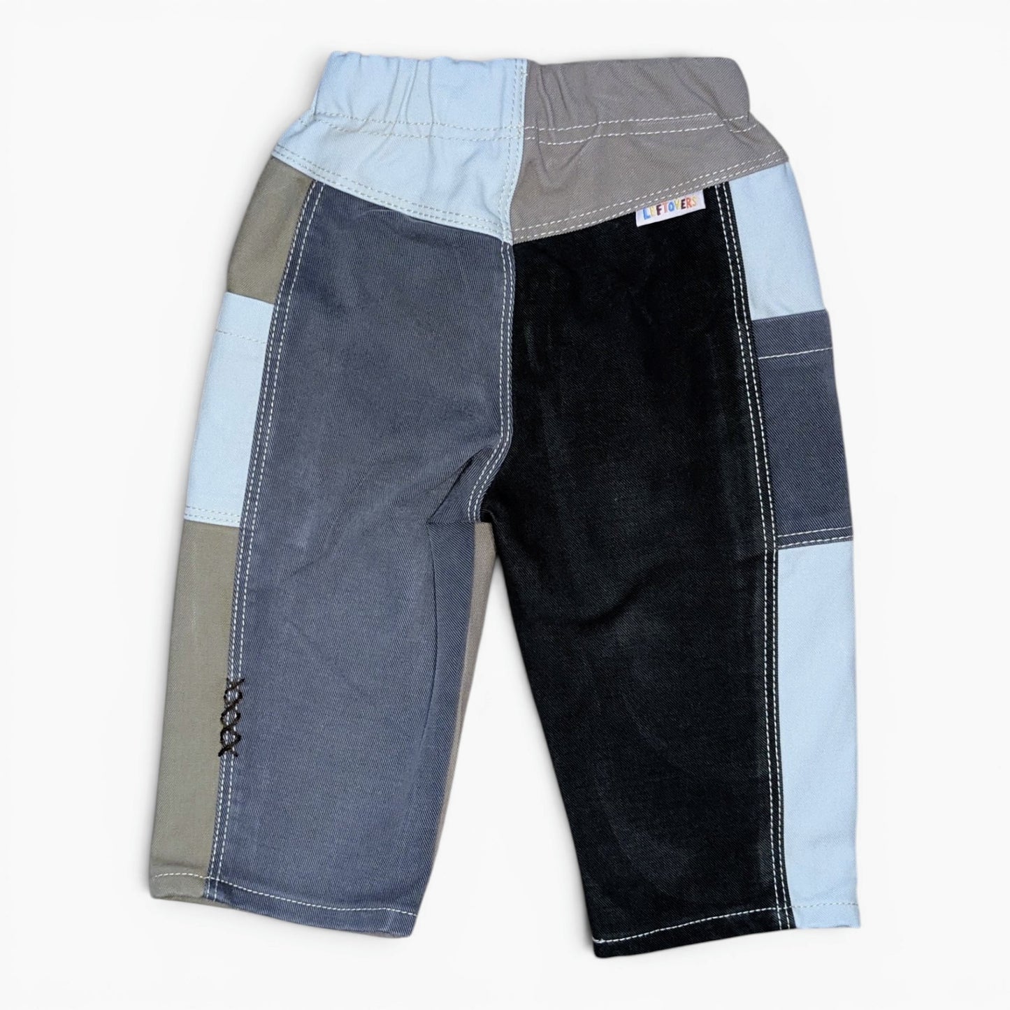 A pair of patchwork color-block pants for kids, featuring a mix of light blue, charcoal gray, slate blue, and black fabrics with contrast white stitching, shown against a plain white background.