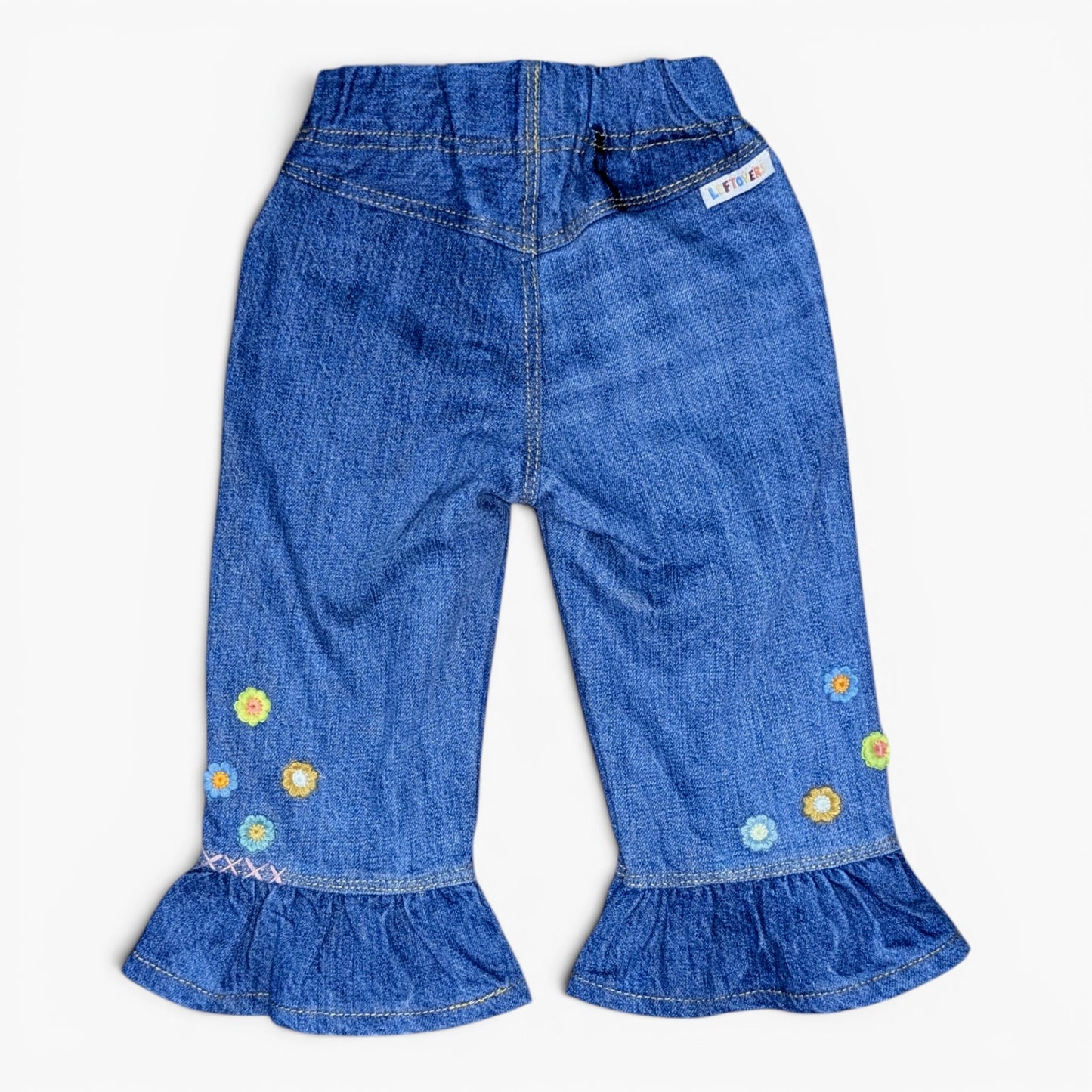 Blue denim pants for toddlers, featuring ruffled hems and colorful embroidered flowers on the lower legs, shown against a plain white background.