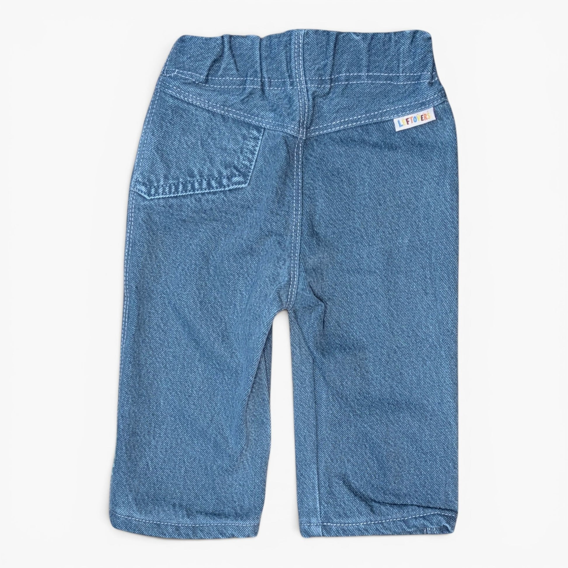 A pair of blue denim pants shown from the back, featuring a wide elastic waistband, a back pocket, and a small white brand label on the upper right side, photographed against a clean white background.