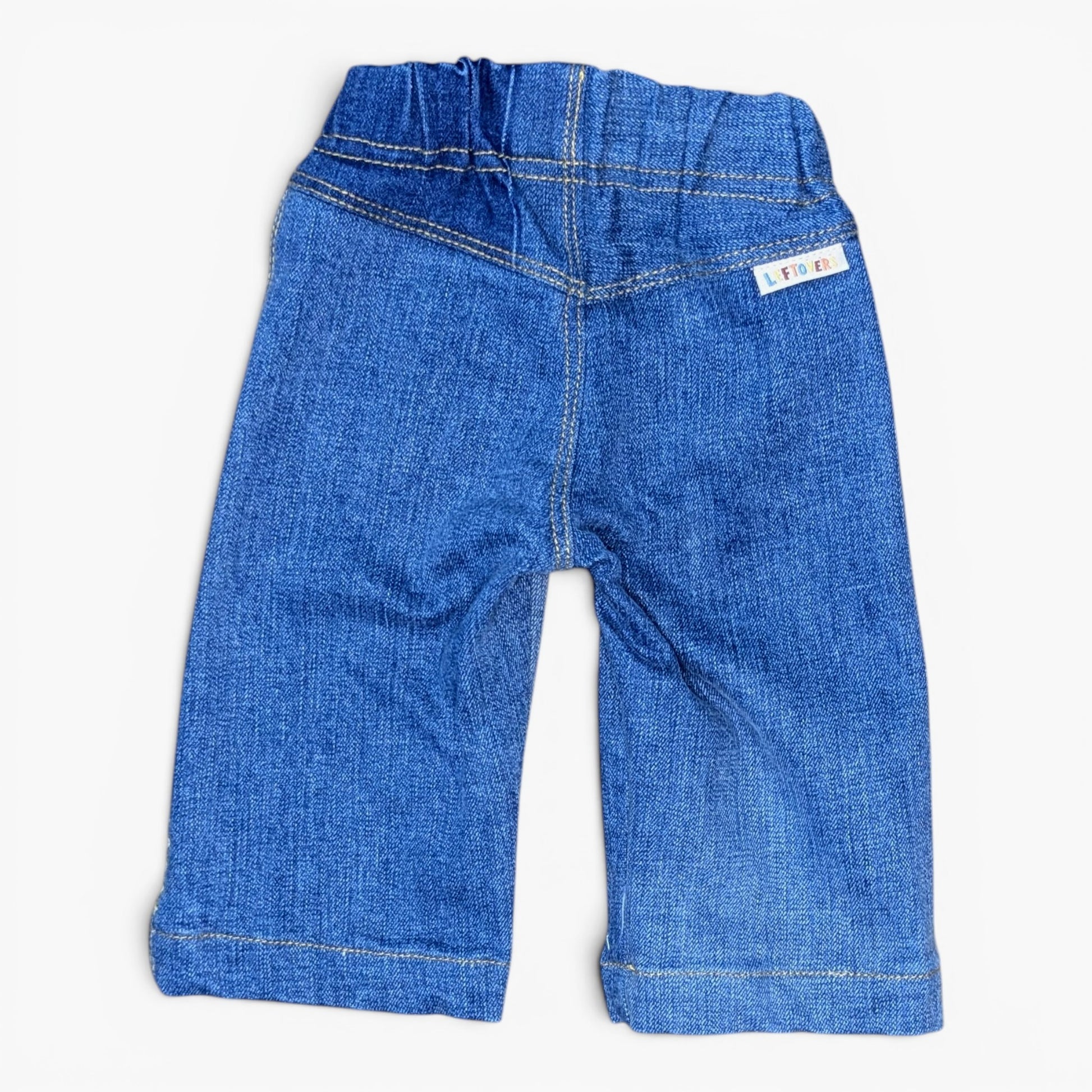 A pair of blue denim jeans for toddlers, shown from the back on a light off-white background. The jeans feature an elastic waistband, classic denim stitching, and a small white brand label on the upper right side.
