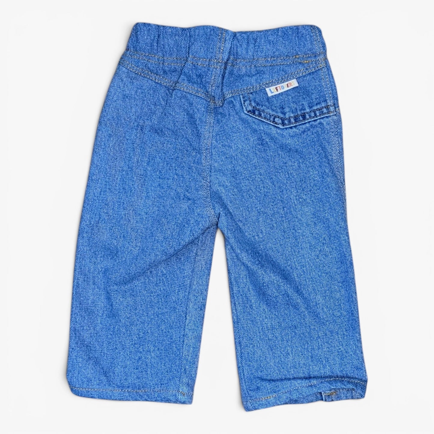 A pair of blue denim pants for toddlers, shown from the back on a light off-white background. The shorts feature an elastic waistband, a back pocket with a small white brand label, and a classic denim texture.