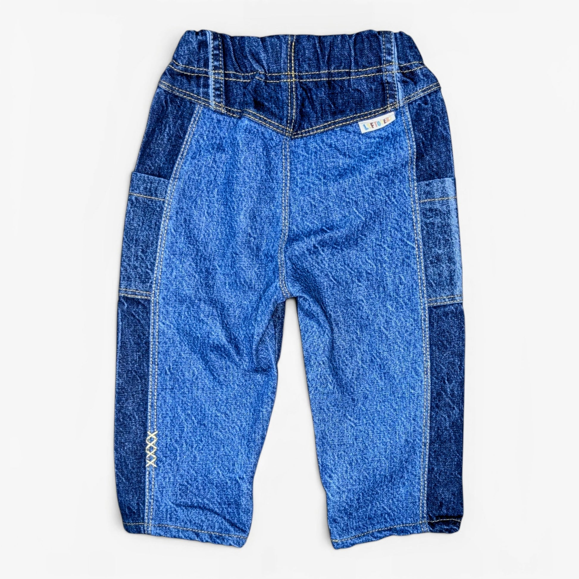 A pair of toddler denim pants featuring a lighter blue main body with darker blue patchwork side panels and pocket details, laid flat on a light gray background.