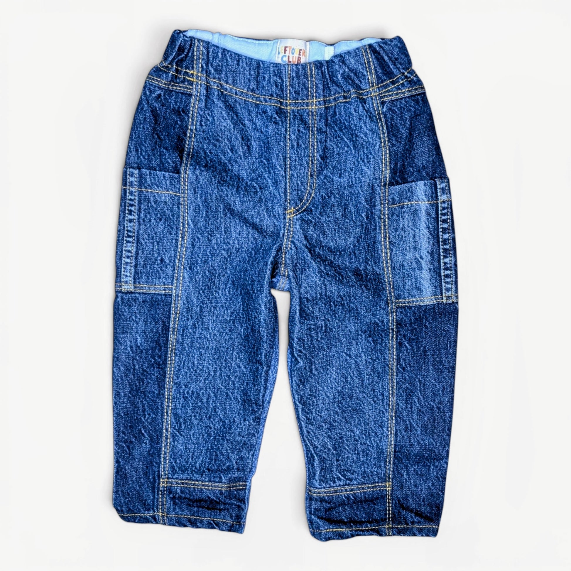 A pair of blue denim pants for toddlers, featuring a wide elastic waistband, contrast yellow stitching, and large patch pockets on the thighs, laid flat against a white background.