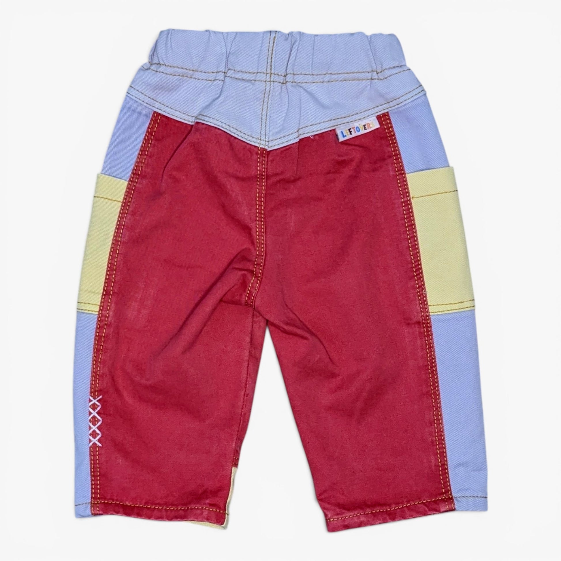 A pair of children's patchwork pants in red, light blue, and pale yellow, laid flat against a white background. The pants feature a red main body, light blue waistband and side panels, and pale yellow color-blocked sections.