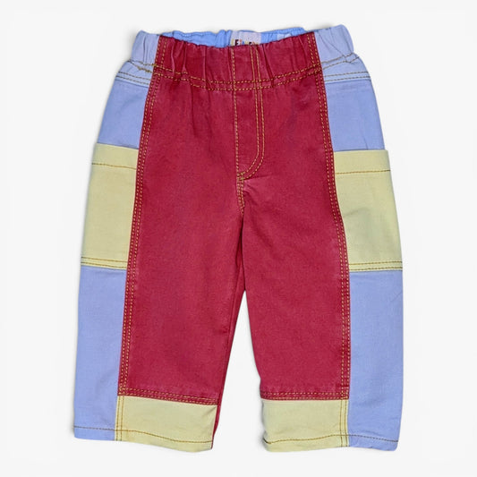 A pair of children's patchwork pants featuring a central red panel, light grey side panels, and pale yellow horizontal stripes, laid flat against a white background.