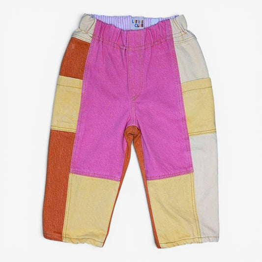A pair of children's patchwork pants in vibrant colors, featuring a large pink panel on the front, a cream panel on the right leg, an orange panel on the left leg, and yellow panels on the upper thighs, laid flat against a light gray background.
