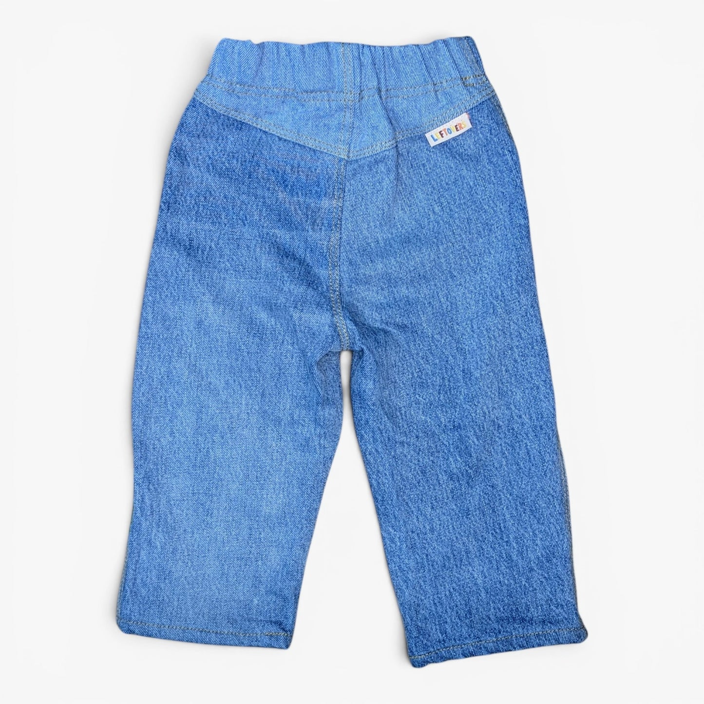 A pair of blue denim pants for toddlers, shown from the back on a light gray background. The back features a wide elastic waistband, a V-shaped back seam, and a small white brand tag on the upper right side.