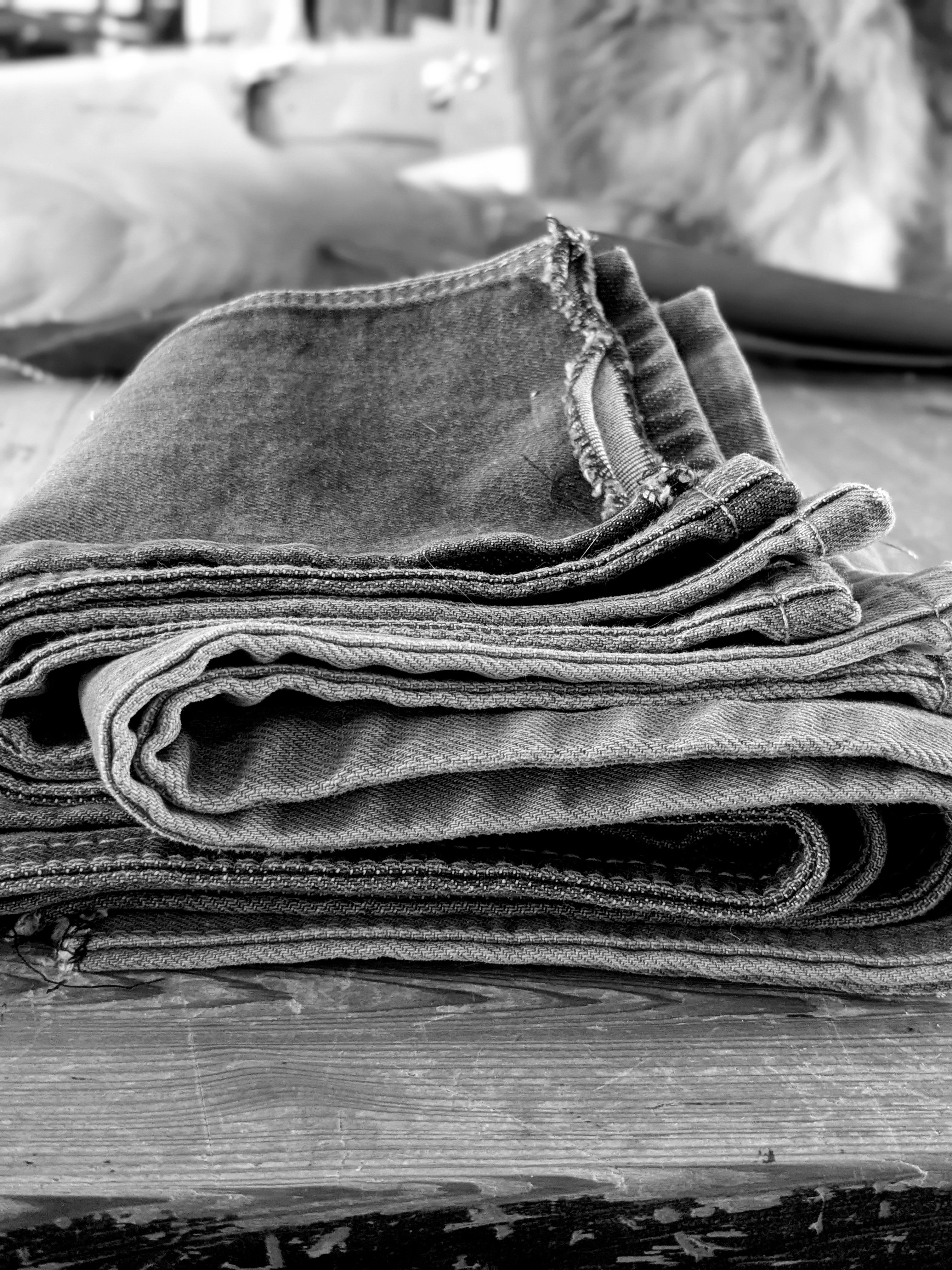 A close-up black and white photograph of folded denim jeans stacked on a rustic wooden surface, with a softly blurred background.