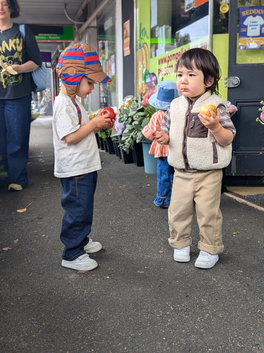 Two toddlers standing outdoors on a street or market walkway, each holding a small fruit. One child wears a white shirt, blue Leftovers Club Stevie Straight jeans, and a brown-and-red striped hat, while the other wears a cream fleece vest over a brown top, tan Leftovers Club Stevie Straight jeans, and white sneakers.