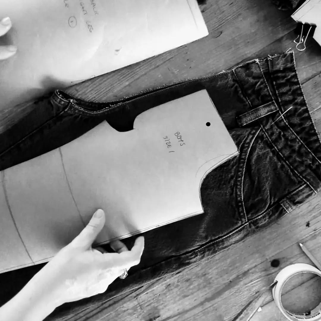 A hand holds a paper garment pattern shaped over dark denim jeans on a wooden work surface, with sewing tools and fabric visible around it.