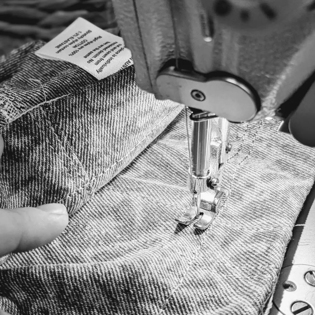 A close-up black and white photo of a sewing machine needle stitching denim fabric for a pair of Leftovers Club Stevie Straight jeans.