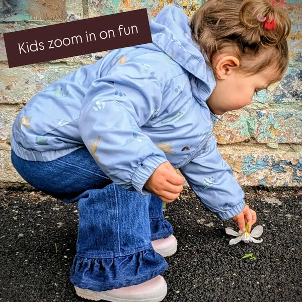 A toddler crouches outdoors to play with a flower on dark asphalt near a weathered stone wall, wearing a light blue hooded jacket with small animal prints, blue jeans, and pink shoes. A brown text banner in the top left reads, 'Kids zoom in on fun'.